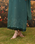 A person outdoors wearing a long teal-green dress or tunic with delicate embroidered motifs and a scalloped lace hem, paired with tan heeled sandals on grass in front of a blurred floral background.