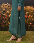 A person wearing a long teal green embroidered kurta with matching pants and tan strappy sandals, standing on grass in front of a floral backdrop.