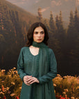 A woman wearing a dark green embroidered kurta with a matching scarf stands in front of a scenic backdrop of mountains, pine trees, and wildflowers.