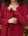 A woman wearing a long-sleeved deep red dress with intricate lace trim, tassel ties, and embroidered floral details, shown in a close-up shot against a blurred outdoor background.