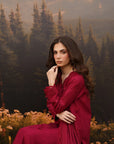 A woman wearing a deep burgundy red embroidered dress sits in front of a forest and mountain backdrop. The dress has long sleeves with lace detailing and a flowing skirt.