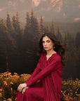 A woman wearing a deep magenta-red embroidered kurta sits in a field of wildflowers with a large scenic backdrop of a pine forest and mountains.