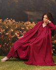 A woman wearing a flowing deep burgundy robe or nightgown sits barefoot on grass in front of a floral backdrop. The garment has a loose, draped silhouette with long sleeves and a deep V-neckline.