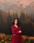 A woman stands in a mountain meadow wearing a long burgundy wrap dress, with a large scenic backdrop of pine trees and snow-capped mountains behind her.