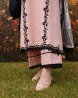 A close-up of a person wearing a light pink traditional outfit with black floral embroidery and lace trim, standing on green grass in front of a blurred floral backdrop.