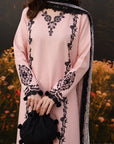 A woman wearing a light pink traditional outfit with black floral embroidery and a black-and-silver patterned dupatta, holding a small black embroidered pouch bag in front of a blurred garden background.