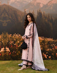 A woman stands outdoors in front of mountains and flowerbeds wearing a light pink traditional outfit with black lace trim and a white printed dupatta. She holds a small black handbag.