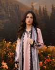 A woman stands outdoors in front of a forested mountain backdrop, wearing a light pink traditional outfit with black embroidered detailing and a sheer gray patterned shawl draped over her shoulders.