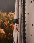 A close-up of a grey traditional garment with black embroidered floral motifs and a black lace-trimmed border, shown on a model against a blurred garden background.