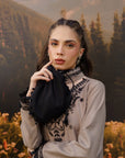 A woman wearing a grey embroidered long-sleeve top with black lace trim, holding a black drawstring pouch against a scenic mountain and forest backdrop.