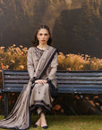A woman sits on a dark wooden bench in front of a floral and forest backdrop, wearing a traditional outfit with a light gray kurta and a matching draped scarf with black embroidery.
