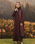 A woman modeling a traditional dark brown salwar kameez set with a matching patterned dupatta, standing in front of a scenic backdrop of mountains, pine trees, and a flower field.
