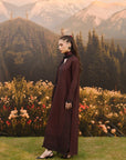 A woman stands in profile outdoors in front of a mountain-and-flower backdrop, wearing a long dark burgundy maxi dress with a high neck and side slit.