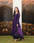 A woman models a deep purple long kurta with matching pants. The kurta features a solid purple upper portion and a sheer black lower skirt panel decorated with purple floral embroidery.