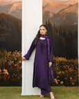 A woman models a deep purple traditional South Asian outfit with matching pants and dupatta, standing next to a white column in front of a scenic backdrop of mountains and flowers.