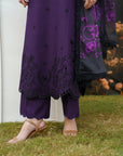 A person outdoors wearing a purple traditional outfit with black embroidered detailing, matching purple pants, and a sheer black dupatta with purple floral embroidery, standing on grass.