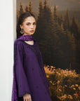 A woman models a deep purple traditional outfit with black embroidered details and a matching purple patterned dupatta, posed against a scenic forest mural background.