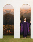 A woman stands between two large arched scenic panels, wearing a deep purple traditional outfit with patterned sleeves. The setting includes green grass at the bottom and a white architectural backdrop behind her.