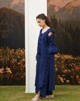 A woman models a deep navy blue embroidered kurta set with matching trousers and a matching handbag, standing outdoors in front of a scenic mountain backdrop.