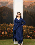 A woman stands outdoors in front of a scenic mountain backdrop and flowerbed, wearing a deep navy blue traditional outfit with a draped shawl and matching pants. A large white archway is behind her.