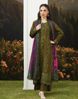 Woman wearing a dark olive green embroidered linen suit with matching dupatta and contrast border, posing in a garden setting with autumn flowers and painted mountain backdrop — Beyond East Winter Tale collection