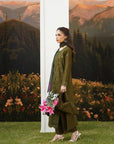 A woman models an olive green traditional outfit with a matching scarf and dark trousers, standing in front of a scenic backdrop of mountains, trees, and flowers.