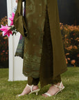 A person outdoors on grass wearing an olive green embroidered traditional outfit with matching pants, white heeled sandals, and holding a matching handbag with pink flowers.