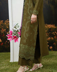 A person outdoors wearing an olive green traditional outfit with matching embroidered kurta and trousers, paired with white heeled sandals. The background shows grass, flowers, and a dark forest mural.