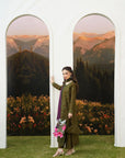 A woman models an olive green traditional outfit with a purple dupatta, standing in front of a white architectural backdrop featuring two arched panels with mountain scenery.