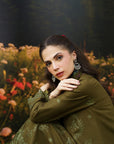 A woman wearing an olive green embroidered traditional outfit with matching embroidered sleeves, posed in front of a blurred outdoor garden background with flowers and trees.