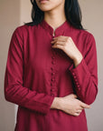 A woman wearing a long-sleeved, deep red button-up tunic with a mandarin collar, shown from the chest down against a neutral beige background.