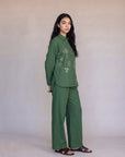 A woman modeling a matching green two-piece set consisting of a long-sleeve embroidered top and wide-leg trousers, standing against a neutral beige studio backdrop.