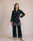 A woman modeling a black two-piece kurta and trouser set with colorful floral embroidery and patterned borders, standing against a solid beige studio backdrop.