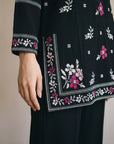 Close-up of a black embroidered tunic or kurta with pink and white floral patterns, shown on a model with a matching black skirt against a plain beige background.