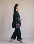 A woman in a black embroidered tunic and wide-leg pants set with floral details, walking in profile against a plain beige studio background.