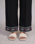Black wide-leg trousers with a decorative embroidered hem featuring white and pink floral patterns, shown on a model wearing off-white quilted slide sandals.