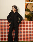 A woman wearing an all-black Pakistani 2 piece co-ord set with a buttoned tunic top and wide-leg trousers, standing against a vintage red-patterned wall — Beyond East Fall Winter 2026 collection