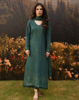 Woman wearing a deep teal green embroidered linen suit with high neckline and matching dupatta, standing in an autumn garden with wildflowers and mountain backdrop — Beyond East Winter Tale collection