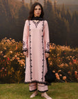 A woman stands outdoors in front of a scenic backdrop of mountains, pine trees, and flowers, wearing a light pink traditional outfit with black embroidered detailing and holding a black handbag.