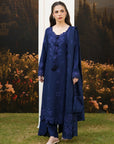 A woman modeling a navy blue traditional outfit with matching embroidered dupatta and trousers, standing outdoors in front of a scenic backdrop of trees and flowers.