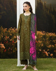 A woman models a three-piece olive green ethnic suit with floral embroidery, matching trousers, and a vibrant magenta and green floral printed dupatta, set against a scenic backdrop of grass, flowers, and a forest.