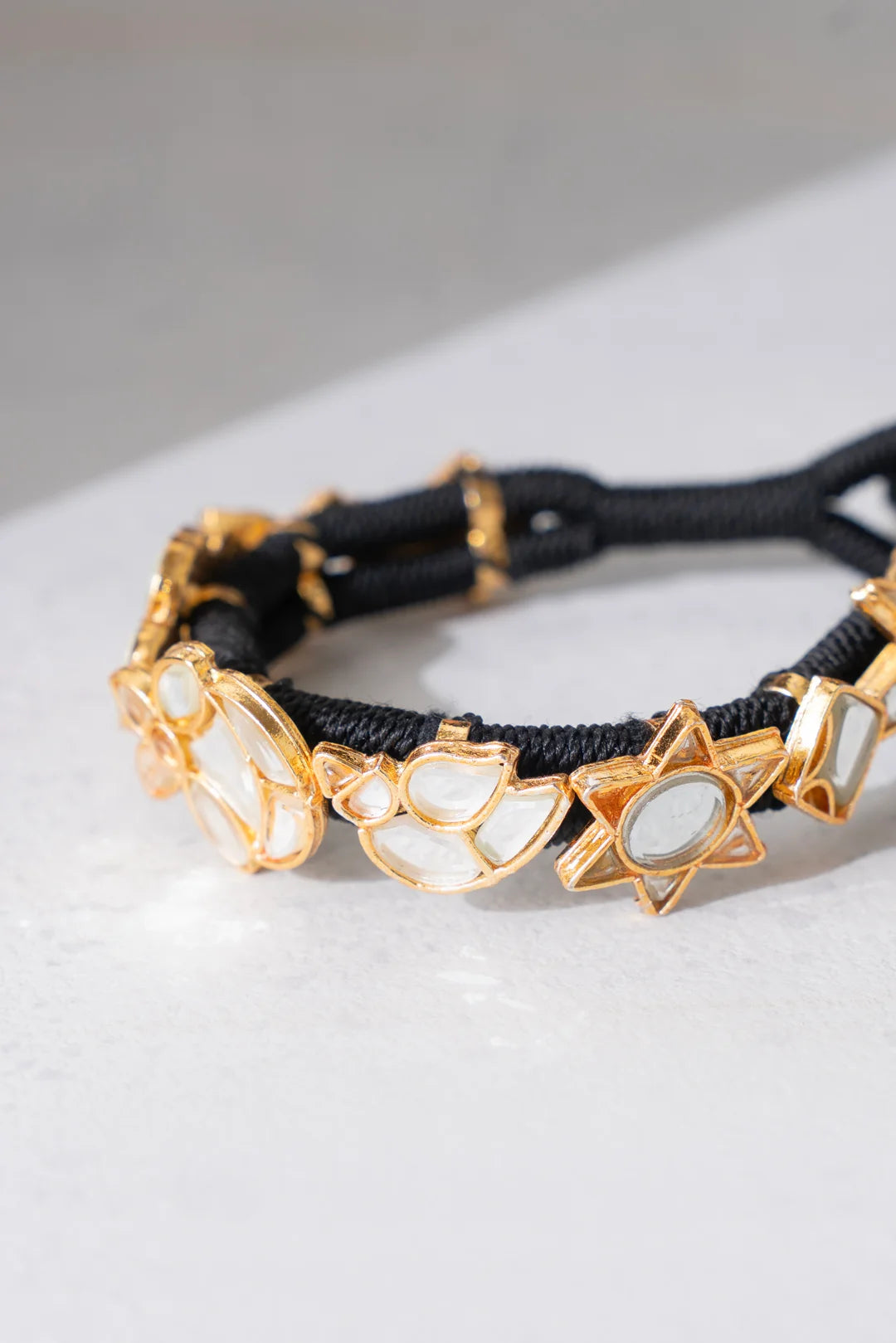 A black braided cord bracelet featuring gold-toned metal charms including crescent moons and six-pointed stars, displayed on a light gray surface.
