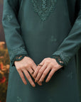 A close-up of a person wearing a dark teal-green embroidered long-sleeve tunic or kurta, with ornate matching embroidery on the chest, cuffs, and scattered small motifs on the fabric. The person's hands are clasped in front, showing rings.
