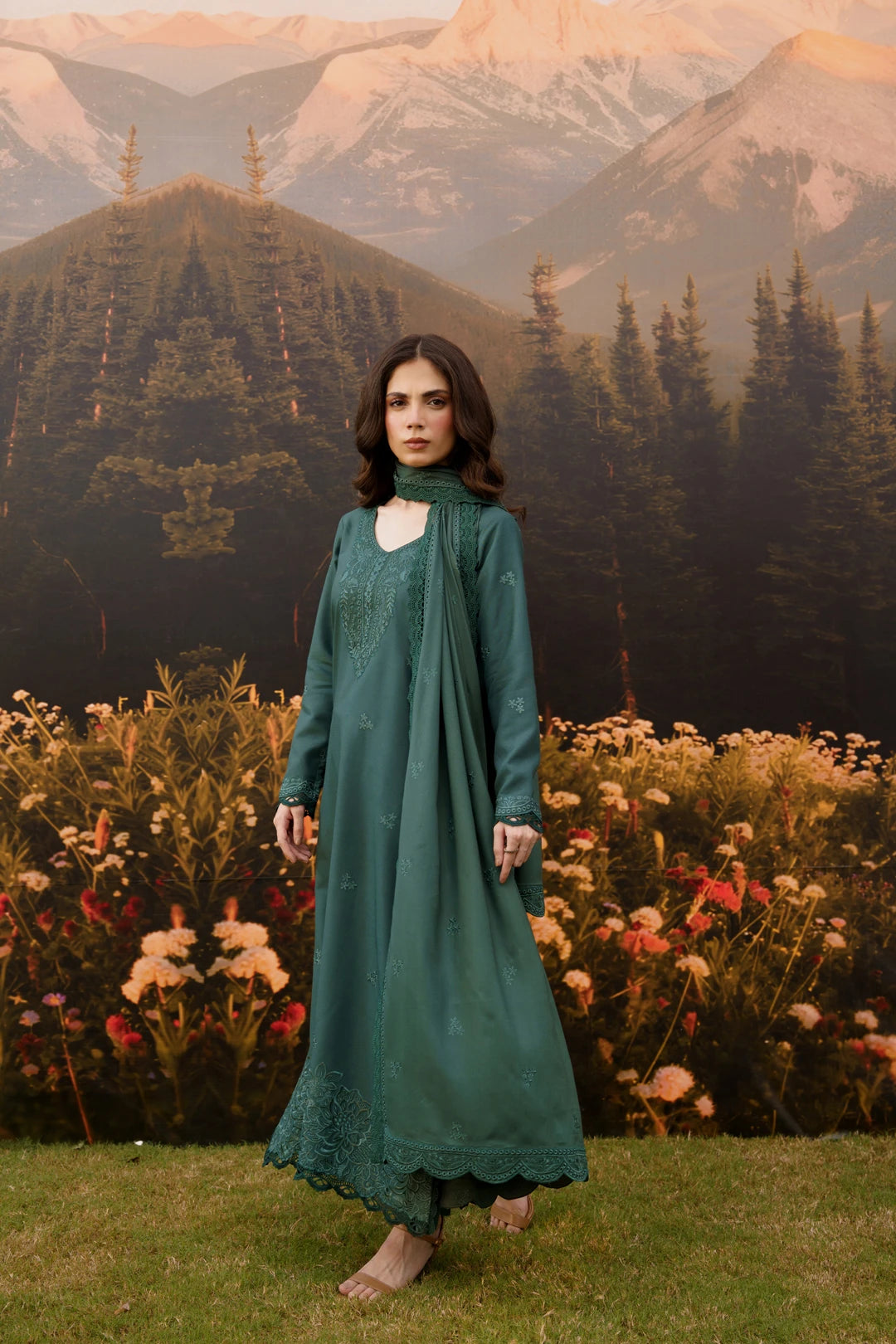 A woman stands outdoors in front of a scenic backdrop of mountains, pine trees, and flowers, wearing a long teal-green embroidered dress with a matching draped scarf.
