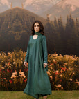 A woman stands outdoors in front of a scenic backdrop of mountains, pine trees, and flowers, wearing a long teal-green embroidered dress with a matching draped scarf.