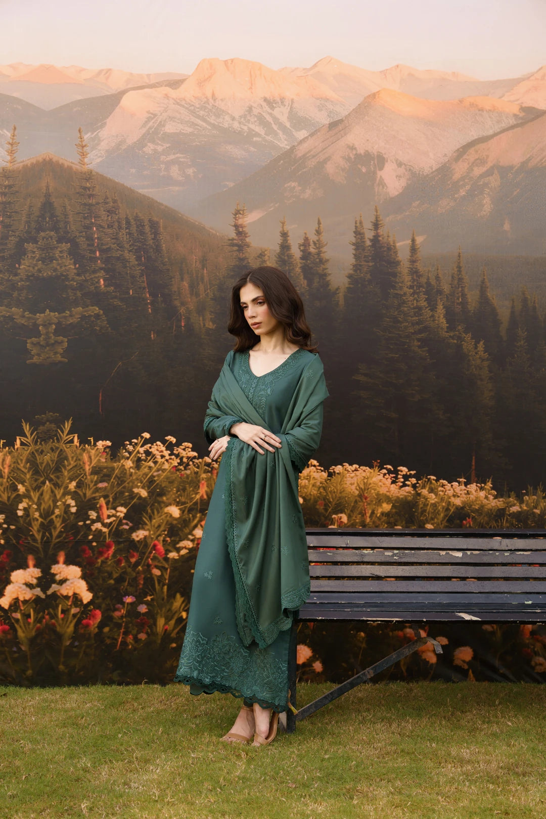 A woman wearing a long, flowing sage green dress stands outdoors in front of a mountain landscape backdrop, with a bench and flowers nearby.