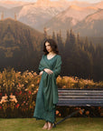 A woman wearing a long, flowing sage green dress stands outdoors in front of a mountain landscape backdrop, with a bench and flowers nearby.