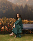 A woman wearing a dark green draped dress and shawl sits on a wooden bench in front of a scenic backdrop depicting mountains, pine trees, and a flowered meadow.