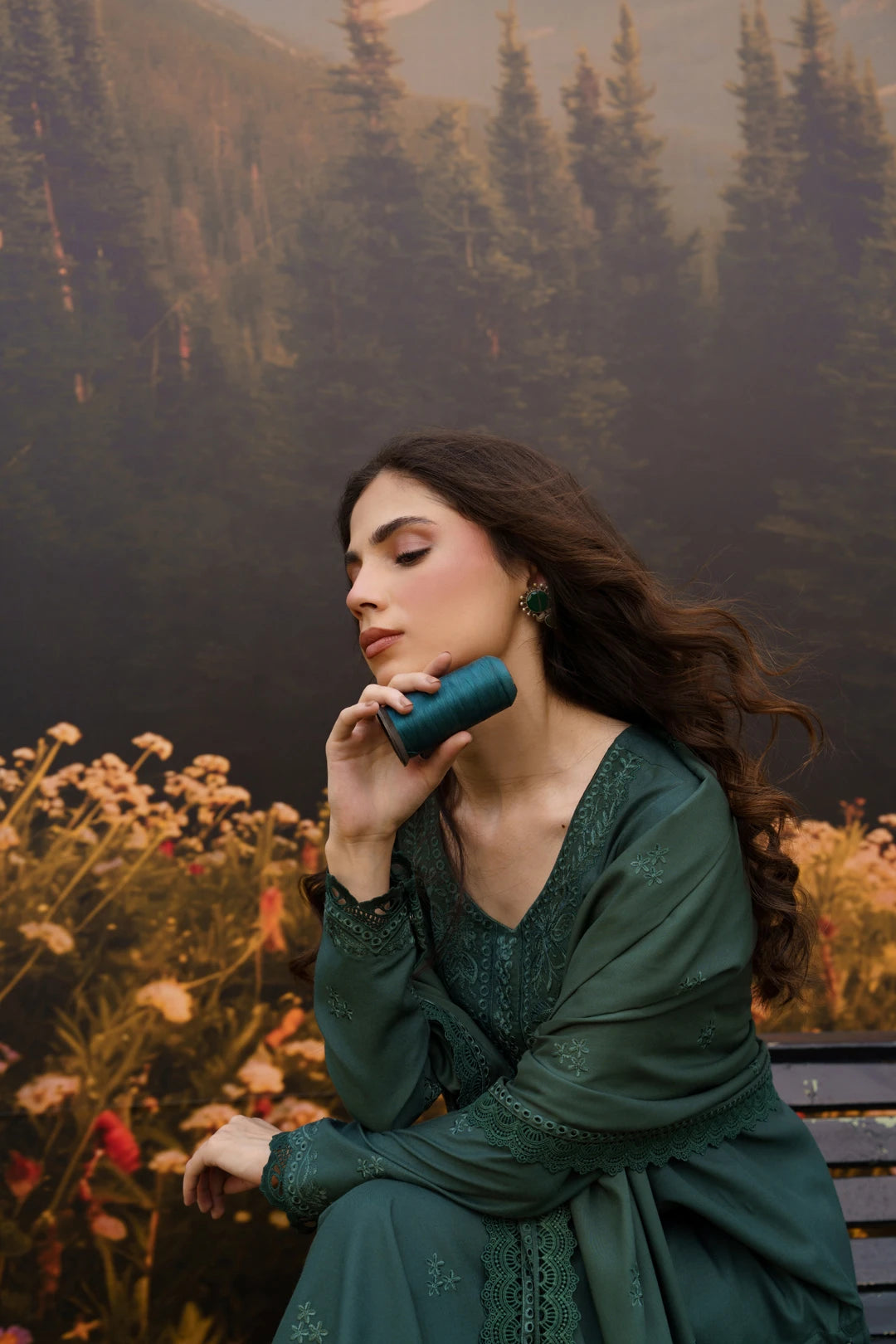 A woman in a dark teal green embroidered dress sits on a bench in front of a forest backdrop, holding a matching teal cylindrical container to her lips.
