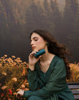 A woman in a dark teal green embroidered dress sits on a bench in front of a forest backdrop, holding a matching teal cylindrical container to her lips.